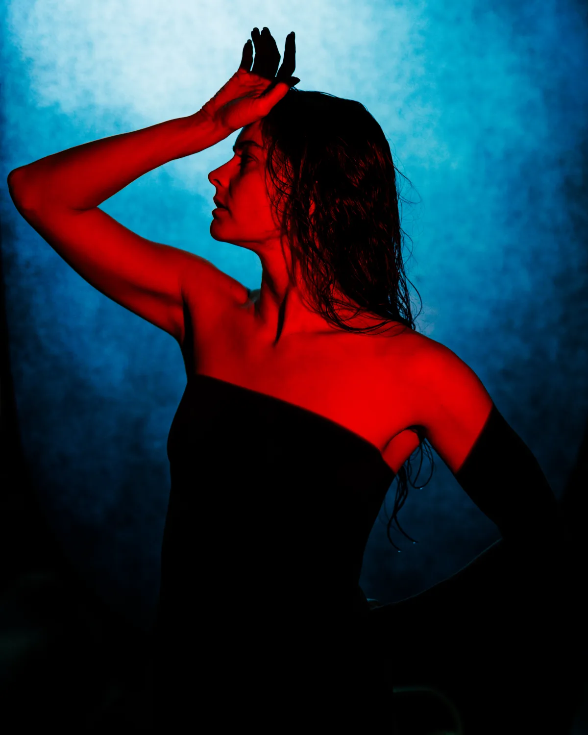Profile portrait lit by a hard red light against a blue backdrop, with most of the body falling into black