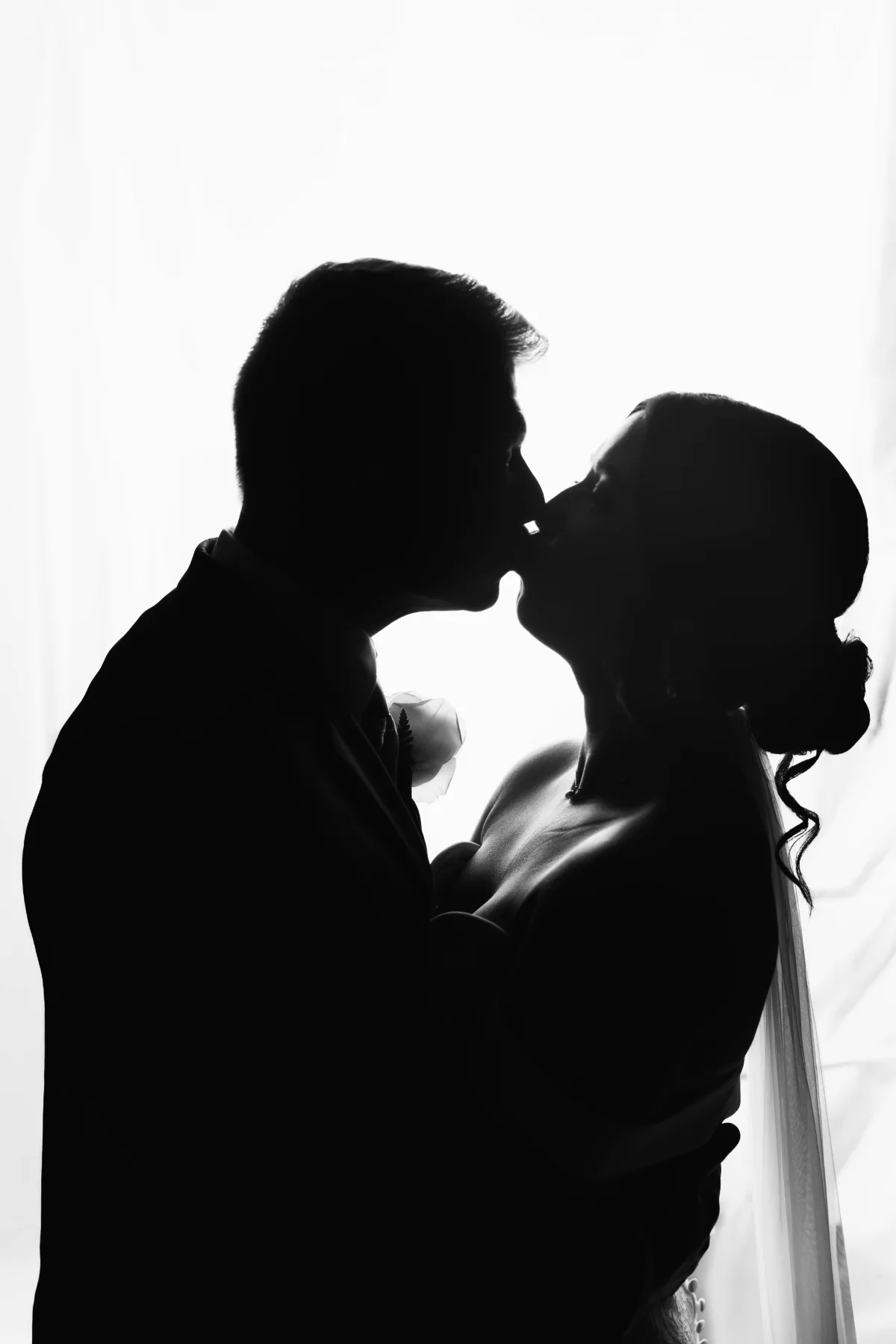 Black-and-white wedding photo of a couple kissing, silhouetted against a bright, soft backlight